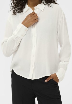 Personne portant une chemise blanche à manches longues boutonnée et un pantalon noir, une main touchant le col de la chemise et l'autre dans une poche.