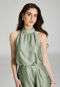 Top halter en satin de couleur verte douce avec un décolleté froncé et une taille élastique. Le mannequin porte des boucles d'oreilles en argent en forme de cercle, mettant en valeur une texture lisse.