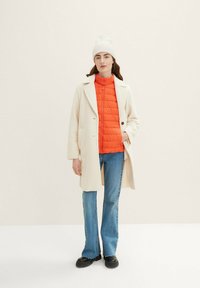 Schicht-Outfit mit einem weißen, strukturierten Mantel, einer leuchtend orangefarbenen Daunenjacke, blauen Jeans und schwarzen klobigen Stiefeln. Weiße Strickmütze.