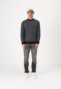 Navy gestreifter Pullover mit langen Ärmeln, gerippten Bündchen und Saum; kombiniert mit grauen Jeans und weißen Sneakers; Model trägt eine hellgraue Kappe.