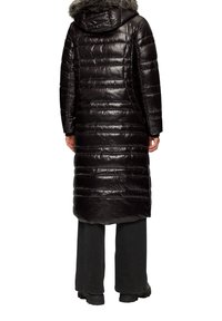 Langer schwarzer Puffer-Mantel mit Kapuze und fellbesatz, in einem gesteppter Design und mit seitlichen Taschen. Getragen mit lockeren schwarzen Hosen und Stiefeln.
