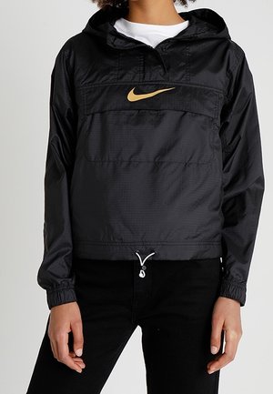Czarna kurtka przeciwdeszczowa Nike z kapturem, złotym logo Swoosh, przednią kieszenią, elastycznymi mankietami i ściągaczem u dołu, noszona na białej koszuli.
