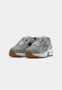 Un paio di scarpe atletiche grigie e bianche con tessuto in rete, lacci bianchi, logo "N" nero e suole in gomma marrone su sfondo bianco.