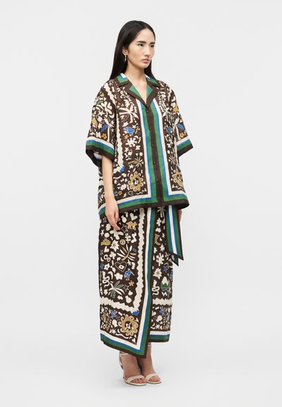 Γυναίκα που φορά έναν φαρδύ καφέ πουκάμισο με κουμπιά και μια αντίστοιχη φούστα μέχρι τους αστραγάλους με πολύχρωμα λουλουδένια και αφηρημένα μοτίβα, στέκεται με ουδέτερη έκφραση.