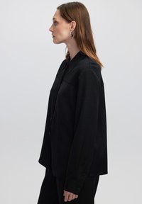 Camicia nera a maniche lunghe con colletto strutturato, caratterizzata da una texture liscia, polsini con bottoni e una vestibilità ampia, mostrata di profilo.