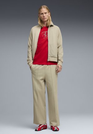 Joven de pie con las manos en los bolsillos, vistiendo un chándal beige, camiseta con el logo rojo de Ferrari y zapatillas rojas y blancas sobre un fondo gris.