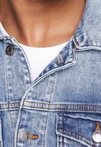 Ljust blå denimjacka med klassisk design, med metallknappar, orange sömmar och framfickor. Närbild av kragen och bröstområdet.