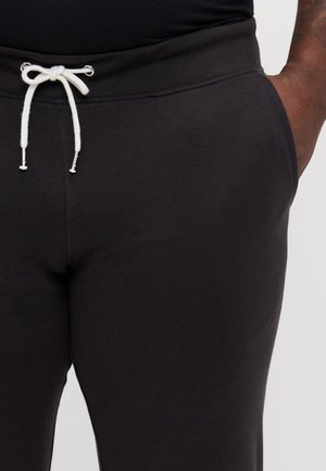 Schwarze Jogginghose mit einem weißen Kordelzug, der zu einer Schleife gebunden ist, getragen von einer Person mit einer Hand lässig in der Tasche.