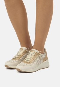 Zapatillas con una combinación de colores beige y crema, que presentan un diseño perforado, acentos texturizados y una suela blanca acolchada. Detalles en hardware dorado.