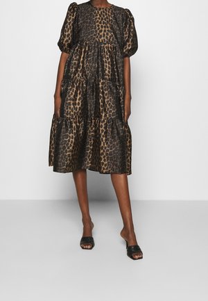 Femme portant une robe à volants marron et noire imprimée léopard avec des manches bouffantes et des talons ouverts noirs, debout sur un fond blanc.