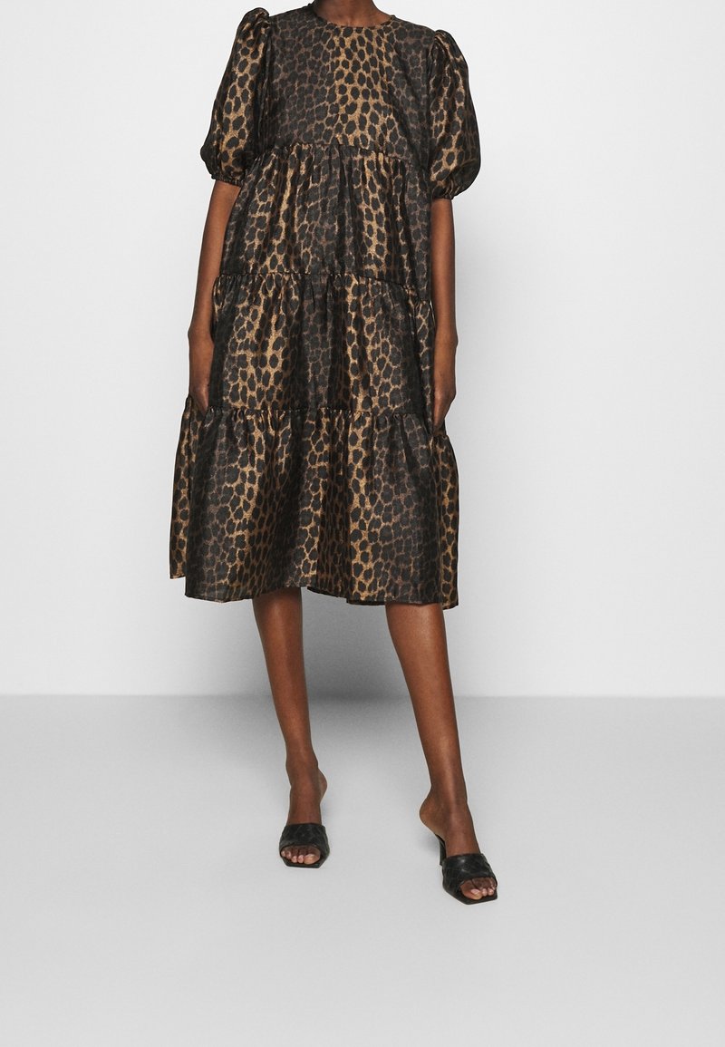 Femme portant une robe à volants marron et noire imprimée léopard avec des manches bouffantes et des talons ouverts noirs, debout sur un fond blanc.