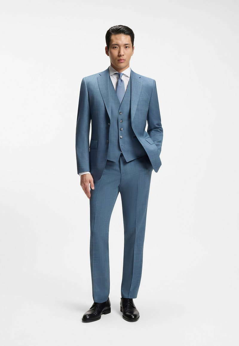 Homme debout en costume trois pièces bleu avec une chemise blanche, une cravate bleue, des chaussures habillées noires et une main dans la poche, sur fond blanc.