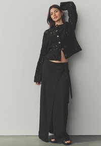 Cardigan negro, recortado, texturizado con recortes intrincados, combinado con una larga falda negra envolvente que presenta una abertura lateral y una cintura atada.