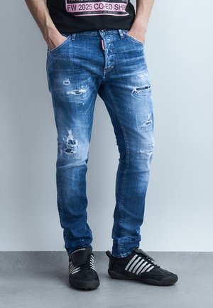 Hombre con jeans azules desgastados y zapatillas negras con rayas blancas, de pie con las manos en los bolsillos sobre un fondo gris.