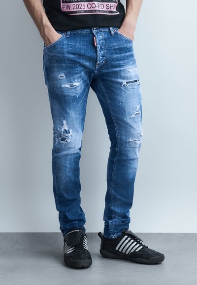 Homme portant un jean bleu déchiré et des baskets noires avec des rayures blanches, debout les mains dans les poches sur un fond gris.