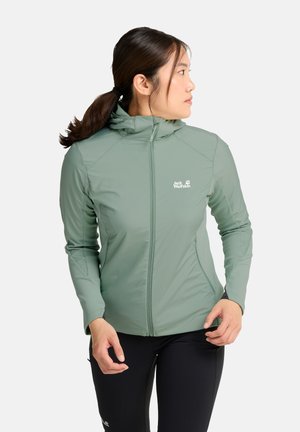 Vrouw met zwarte paardenstaart die een lichtgroene hoodie met rits en een zwarte broek draagt, kijkt naar rechts tegen een witte achtergrond.