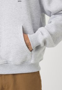 Svijetlosiva dukserica s rebrastim rubovima i manžetama. Ima prednju džepu i minimalistički logo iznad džepa. Mekana, teksturirana tkanina.