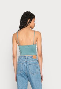 Um top de seda ajustado em cor teal com alças finas, combinado com jeans de ganga azul claro de cintura alta. Os jeans apresentam um patch de couro na parte de trás.