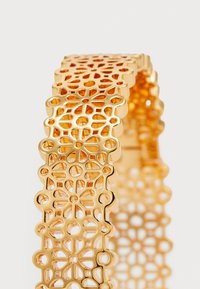 Gullmetall cuff-armbånd med et utskåret geometrisk mønster, med glatte kanter og en teksturert overflate for visuell interesse.