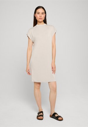 Nő áll, rövid ujjú, bézs, térdig érő ruhát és fekete csatos szandált visel egyszínű szürke háttér előtt.