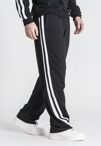 Sorte sweatpants med hvide sidelister, lavet af blødt stof. Har elastisk talje og dybe lommer. Båret med hvide sneakers.