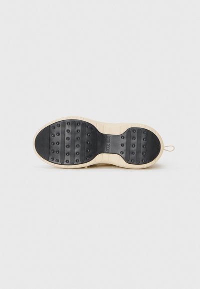 Sole of beige shoe with black tread featuring raised circular grips and horizontal textured lines, set against a plain white background.