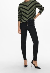 Pull vert olive à rayures chevron noires, coupe décontractée. Jean noir taille haute avec fermeture à cinq boutons. Talons pointus noirs.