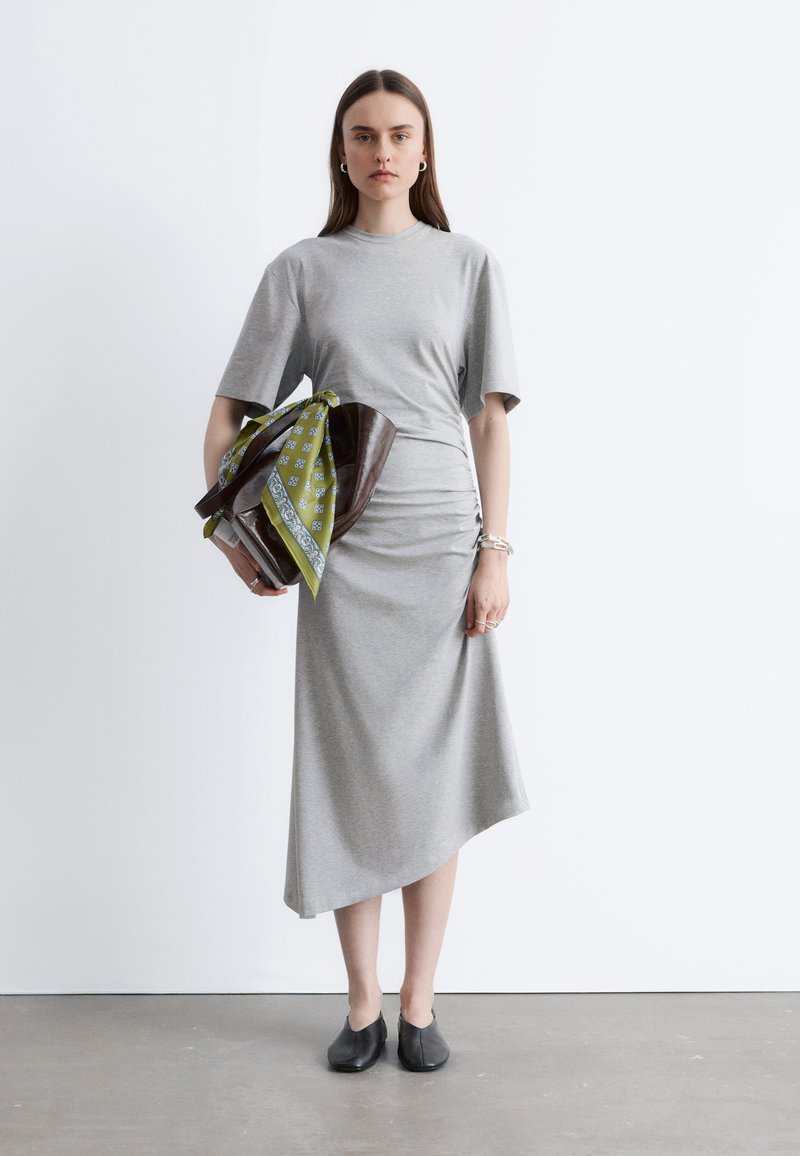 Femme portant une robe midi grise et des chaussures noires, tenant un sac marron avec un foulard à motifs verts, debout sur un sol gris avec un mur blanc en arrière-plan.