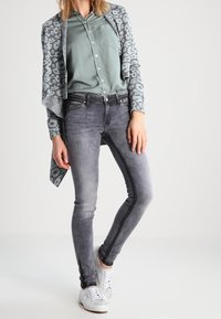 Šedý vzorovaný kardigan přes světle zelenou košili, spárovaný se šedými úzkými džíny a bílými teniskami. Tkaniny jsou hladké a dobře padnou.