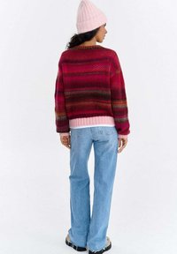 Dziany sweter z wielokolorowym wzorem pasków w odcieniach różu, czerwieni i brązu, z różowym ściągaczem. Połączony z jasnoniebieskimi dzwonowatymi jeansami.