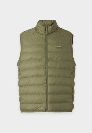 Jeune homme debout portant un gilet matelassé vert olive sur un t-shirt noir à manches longues, un pantalon beige à jambes larges et des baskets blanches.