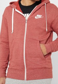 Red Nike zip-up hoodie with a double-layered hood, white drawstrings, kangaroo pockets, and logo on the chest. Cotton blend fabric.