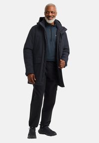 Veste imperméable gris foncé avec capuche, deux poches avant et un devant partiellement zippé. Portée sur une chemise bleue à manches longues et un pantalon noir.