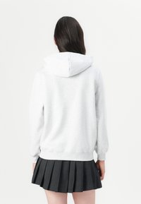 Personne aux longs cheveux foncés portant un sweat à capuche gris clair et une jupe plissée noire, vue de dos sur fond blanc.