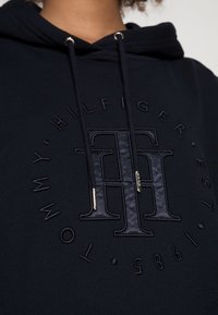 Sudadera negra con un logo en relieve "TH" en el centro. Presenta cordones con puntas doradas y una textura suave. El diseño incluye texto circular.