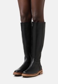 Beine in hohen schwarzen Lederstiefeln mit seitlichem Reißverschluss und braunen Gummisohlen, stehend vor einem weißen Hintergrund.
