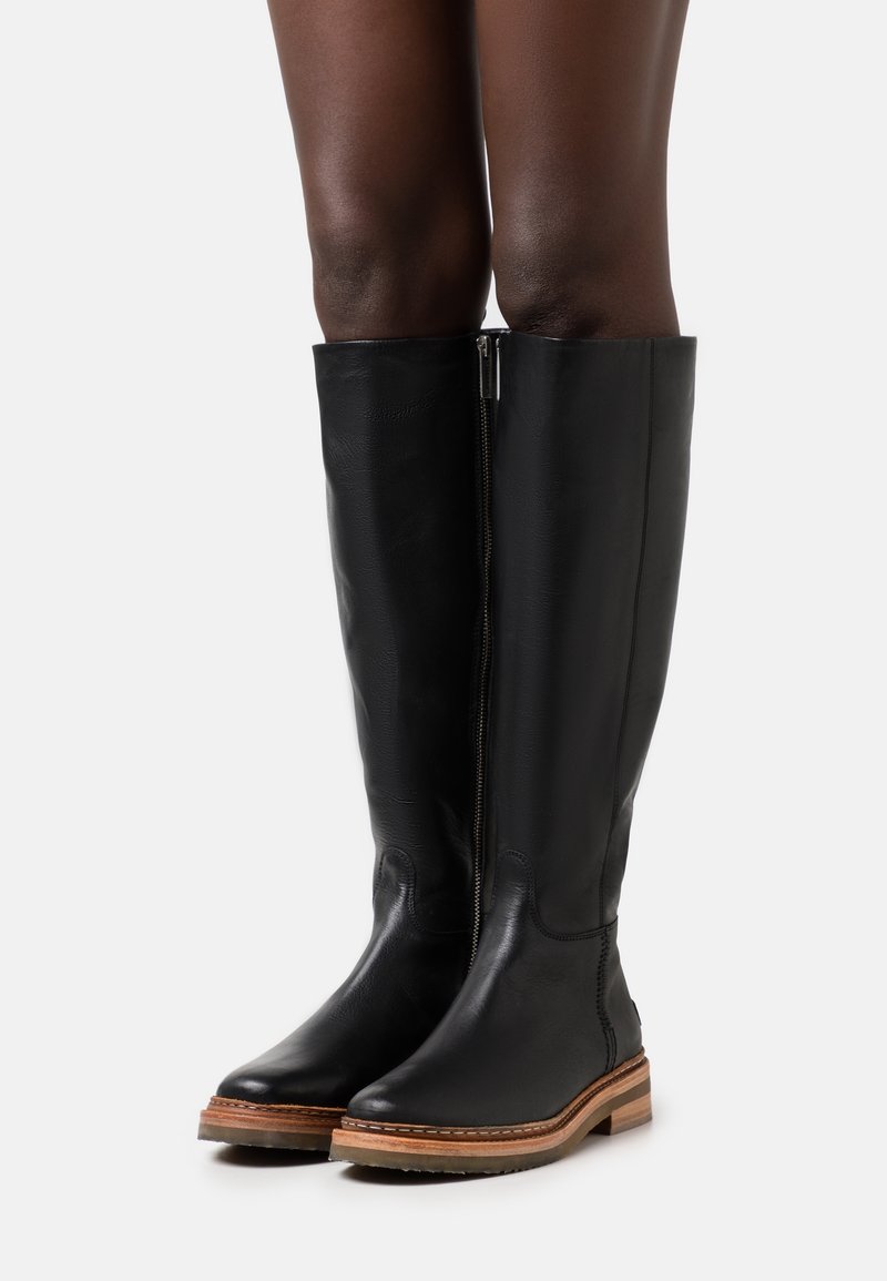 Beine in hohen schwarzen Lederstiefeln mit seitlichem Reißverschluss und braunen Gummisohlen, stehend vor einem weißen Hintergrund.