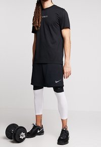 Black athletic t-shirt with logo, paired with black shorts and white compression leggings. Black sneakers and dumbbells are on the floor.