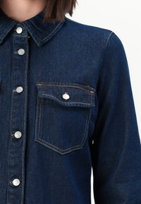 Chemise en jean bleu foncé avec boutons-pression argentés, rabat de poche poitrine boutonné, et coutures contrastantes, portée par une personne aux cheveux courts et bruns.