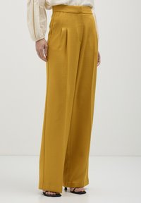 Donna che indossa pantaloni a gamba larga color senape, una blusa crema con maniche a sbuffo e sandali neri con tacco aperto, in piedi su uno sfondo neutro.