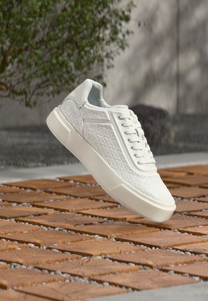 Weißer Sneaker mit geometrischem Muster und dicker Sohle, schwebend über einem Holzgitterboden im Freien, mit Grünpflanzen und Wand im Hintergrund.