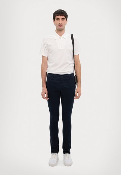 Hvit kortermet polo med glidelåsdetalj, kombinert med marineblå smale bukser og hvite joggesko. Enkelt bakgrunn.