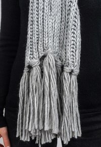 Tjock grå stickad scarf med långa, texturerade fransar. Tillverkad av tjockt garn, har flätade knutar i ändarna. Mjuk och varm i utseendet.