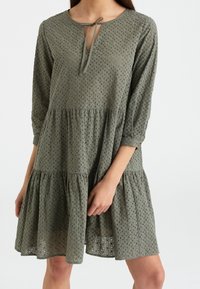 Vestido verde oliva con mangas tres cuartos, diseño de ojal cortado, escote redondo, cierre con lazo y dobladillo con volantes en capas. Tejido suave y texturizado.