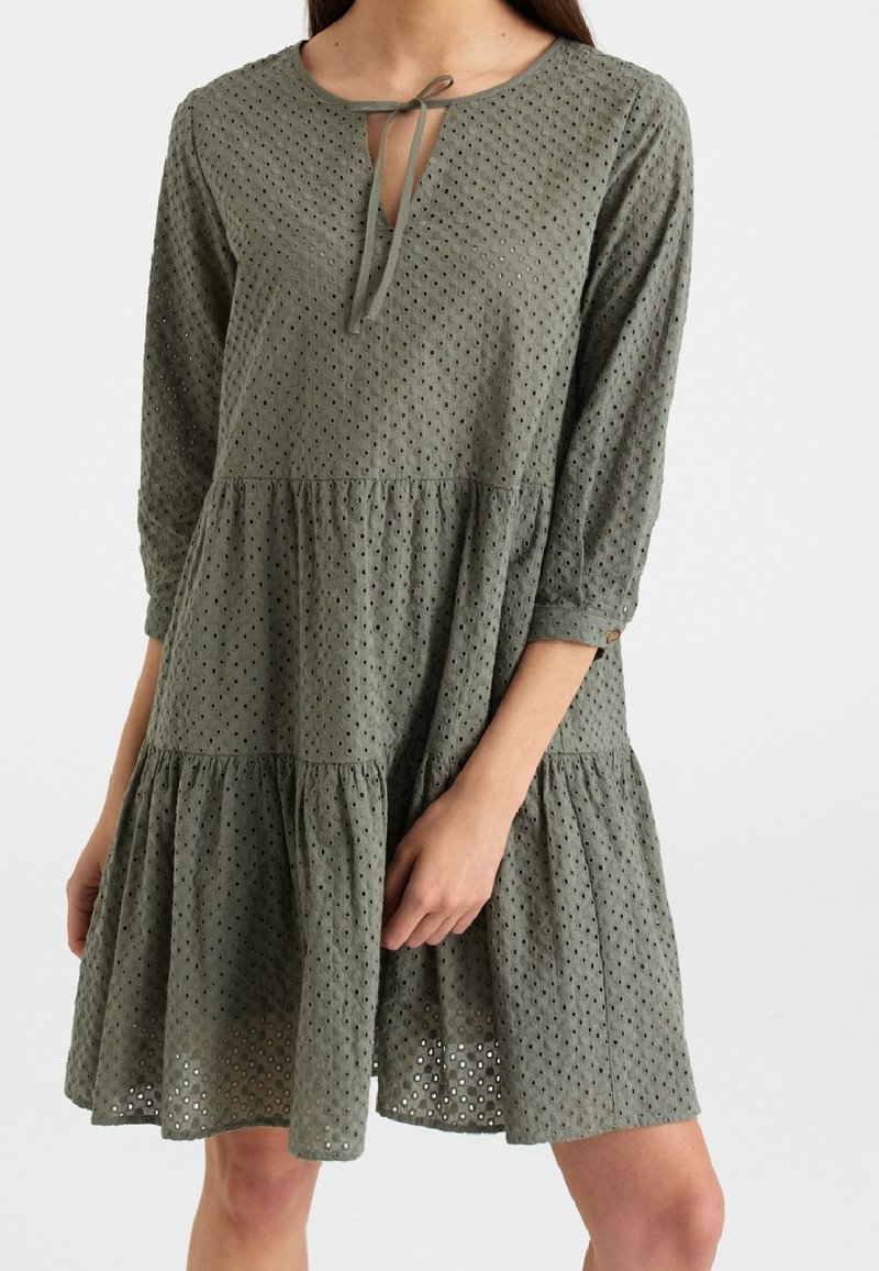 Vestido verde oliva con mangas tres cuartos, diseño de ojal cortado, escote redondo, cierre con lazo y dobladillo con volantes en capas. Tejido suave y texturizado.