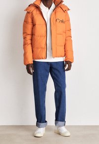 Mężczyzna w pomarańczowej puchowej kurtce, białej koszuli, niebieskich podwiniętych dżinsach i białych trampkach, stoi na tle gładkiej ściany.