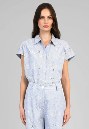 Femme portant un haut bleu clair à manches courtes boutonné et un pantalon taille haute assorti avec une broderie florale blanche.