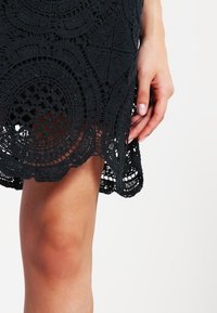 Main reposant à côté de la cuisse vêtue d'une jupe en dentelle noire crochetée avec des motifs circulaires et en forme de losange.