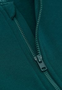 Vêtement zippé vert en tissu lisse et texturé, avec une fermeture éclair visible et des accents côtelés près du col.