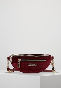 Riñonera de ante burdeos con cremalleras y herrajes en tono dorado, que presenta un logotipo destacado de "GUESS" y detalles de cadena. Dos compartimentos con cremallera.